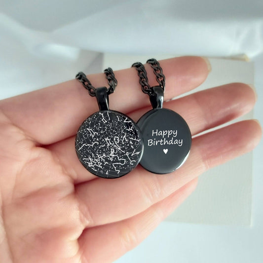 Two black necklaces with engraved star map and text held in a hand against a light background