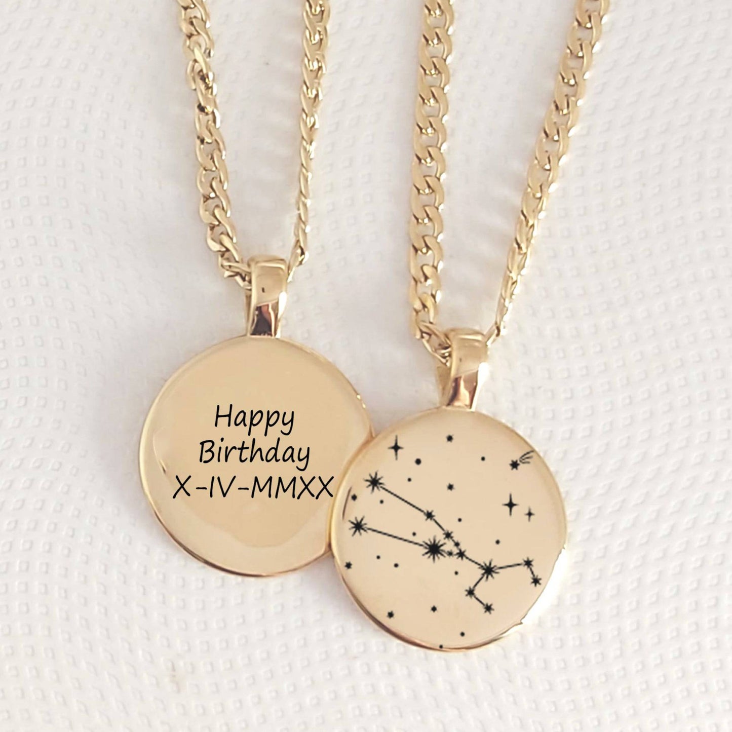 Two gold necklaces with circular pendants, one engraved with 'Happy Birthday' and a date, the other with a constellation design, on a textured white background.