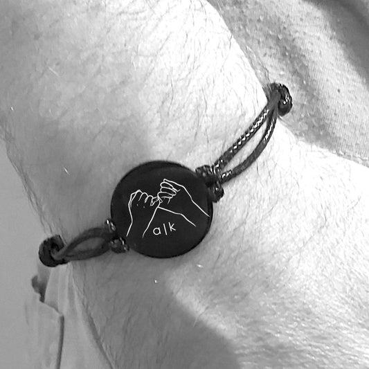 Bracelet with a circular charm featuring a pinky promise sign and two initials, worn on a wrist.