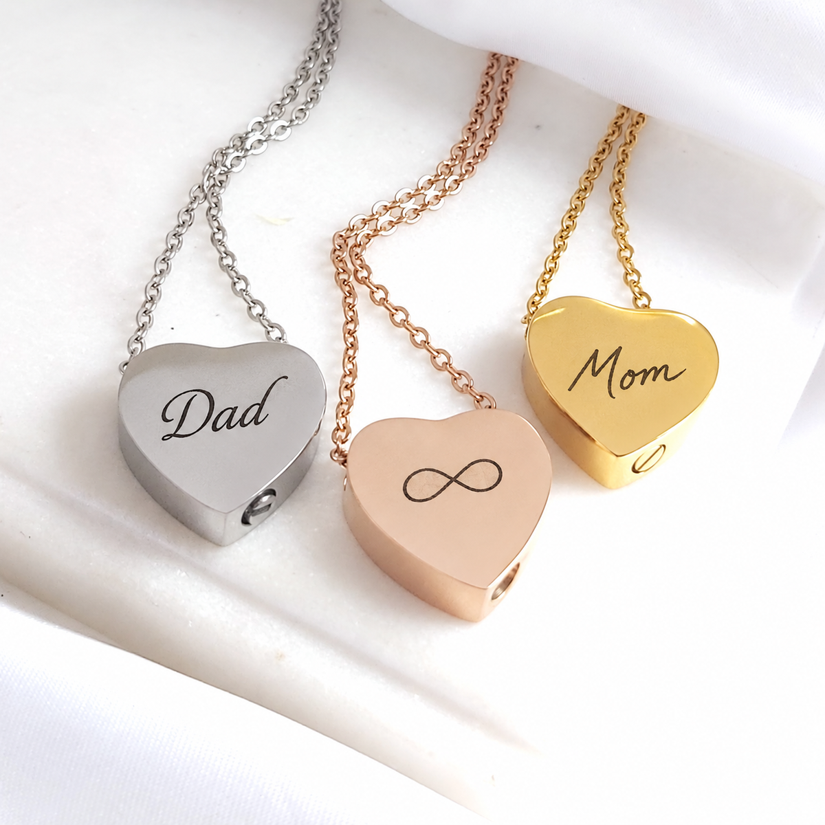 Three polished heart-shaped urn necklaces with matching chains displayed on white fabric. The silver pendant is engraved with “Dad,” the rose-gold-tone pendant features an infinity symbol, and the gold pendant is engraved with “Mom.” Small circular filling openings are visible along the sides of the pendants.