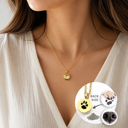 A woman wearing a small round gold necklace with a clear glass centre and delicate gold chain. A circular inset labelled “Back Side” shows the polished back engraved with a black paw print. Close-up images of a paw making a print and a dog’s nose appear beside a small pile of ashes.