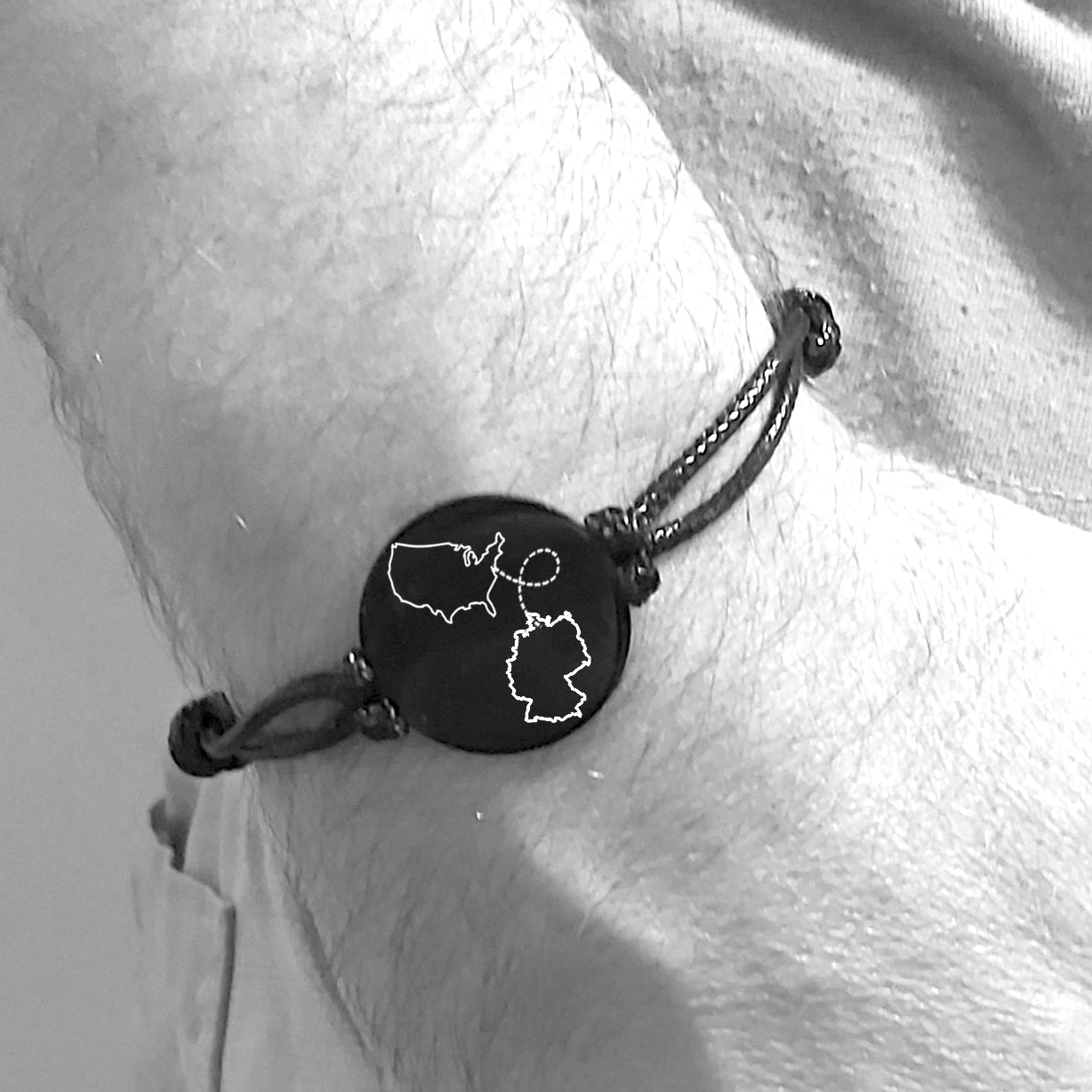 Close-up image of a black leather long-distance bracelet held in both hands, featuring a 20mm engraved coin with two map outlines and a connected, representing a personalized two-locations keepsake.