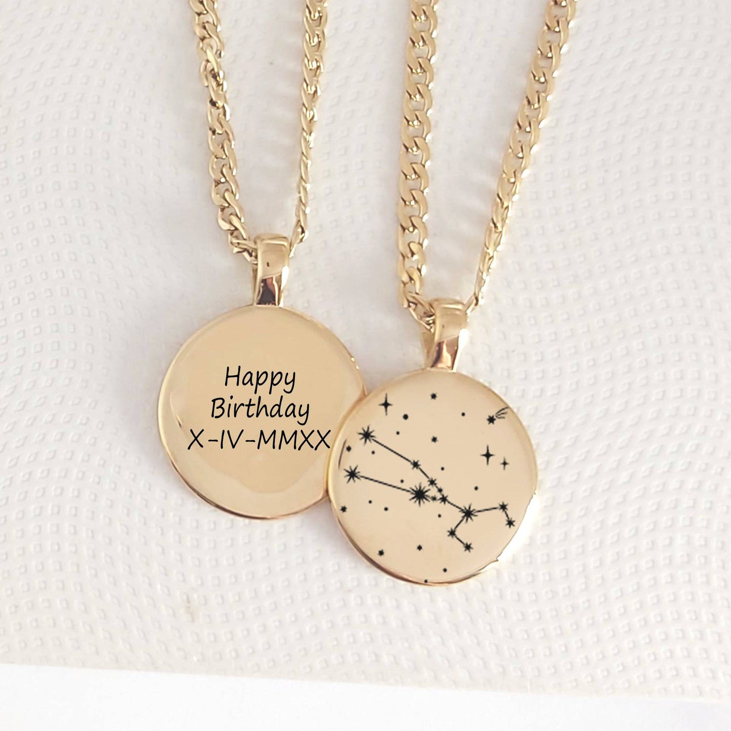 Two gold necklaces with circular pendants, one engraved with 'Happy Birthday' and a date, the other with a constellation design, on a textured white background.