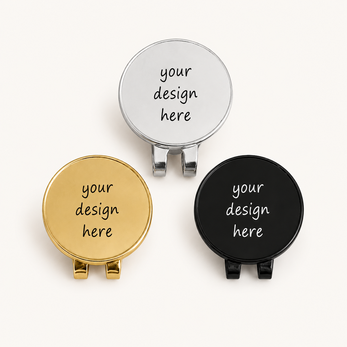 Three round personalized stainless steel golf markers with attached hat clips, displayed in polished silver, gold, and black finishes. Each marker shows the sample engraving “your design here” in a casual handwritten font.