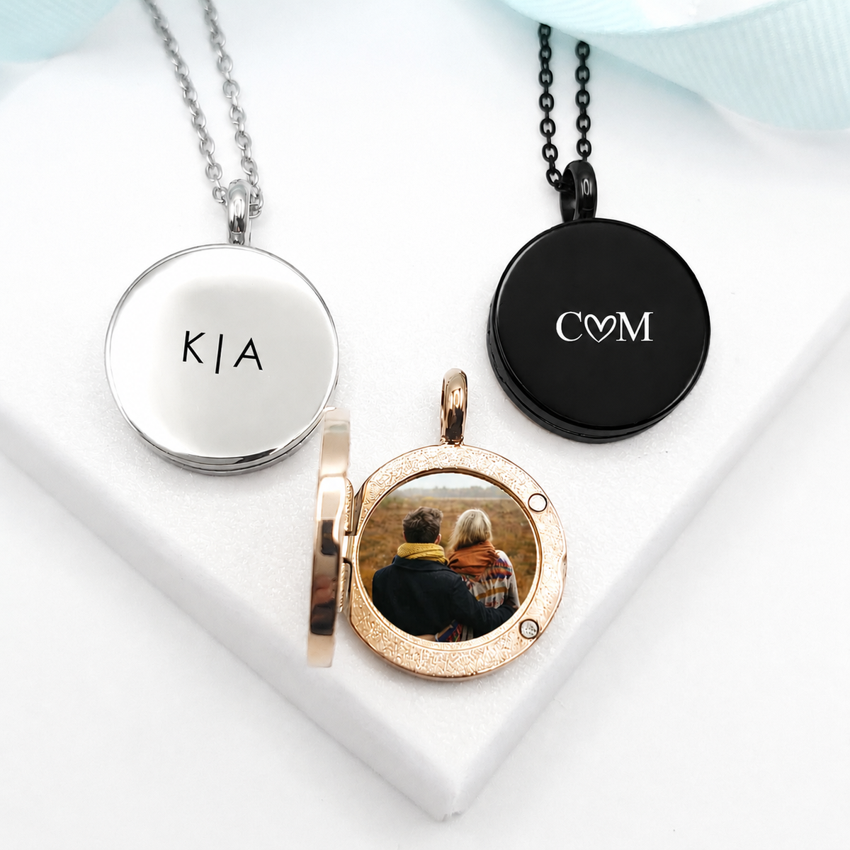 Three round stainless steel photo lockets in polished silver, black, and rose-gold finishes. The closed silver locket is engraved with “K | A,” the black locket features “C♡M,” and the open rose-gold locket displays a photograph of a couple. Each locket hangs from a coordinating chain.