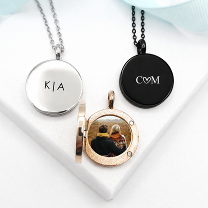 Three round stainless steel photo lockets in polished silver, black, and rose-gold finishes. The closed silver locket is engraved with “K | A,” the black locket features “C♡M,” and the open rose-gold locket displays a photograph of a couple. Each locket hangs from a coordinating chain.