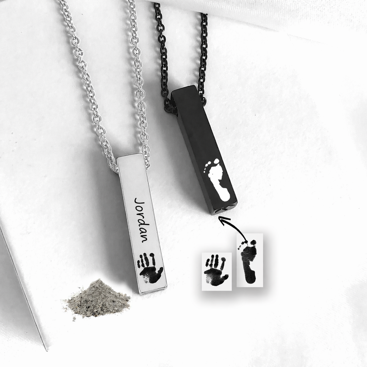 Two personalized baby footprints and handprints necklace pendants shown on chains, one silver-toned with a handprint and the name 'Jordan' and one black-toned with a footprint. Small powder and printed sample cards are included on a white background.