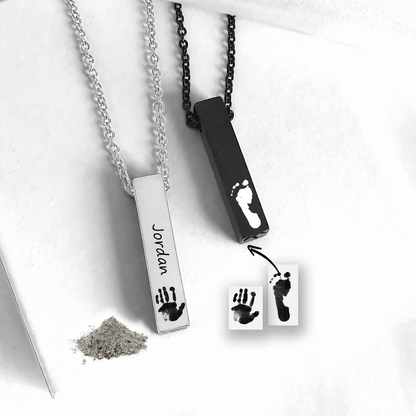Two personalized baby footprints and handprints necklace pendants shown on chains, one silver-toned with a handprint and the name 'Jordan' and one black-toned with a footprint. Small powder and printed sample cards are included on a white background.