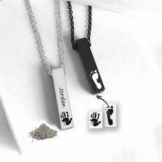 Two personalized baby footprints and handprints necklace pendants shown on chains, one silver-toned with a handprint and the name 'Jordan' and one black-toned with a footprint. Small powder and printed sample cards are included on a white background.