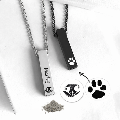 Two rectangular bar urn necklaces on white fabric, one polished silver with “Marley” and a nose print, and one black with a white paw print. Matching silver and black chains are attached. The image also shows circular examples of a pet nose print and paw print, a small pile of ashes, and a metal filling tool.