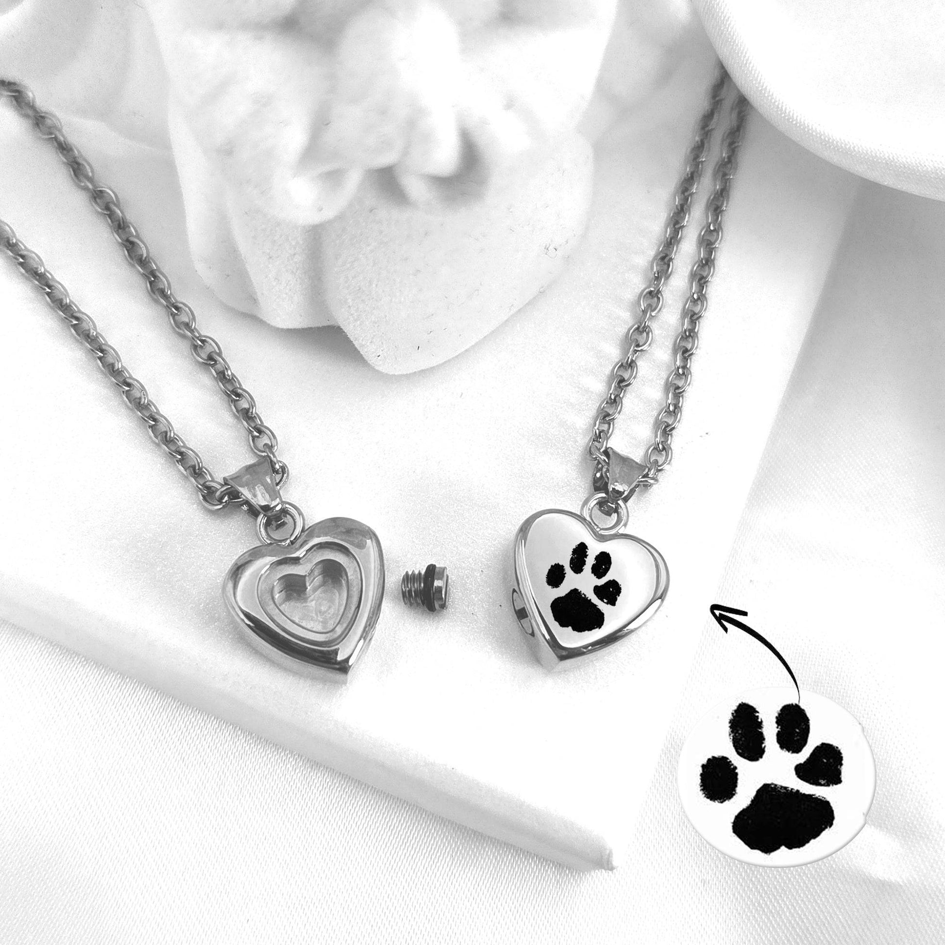 Two silver heart-shaped urn  pendants with paw prints on a white surface.