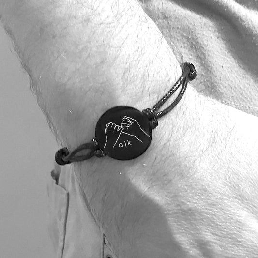 Bracelet with a circular charm featuring a pinky promise sign and two initials, worn on a wrist.