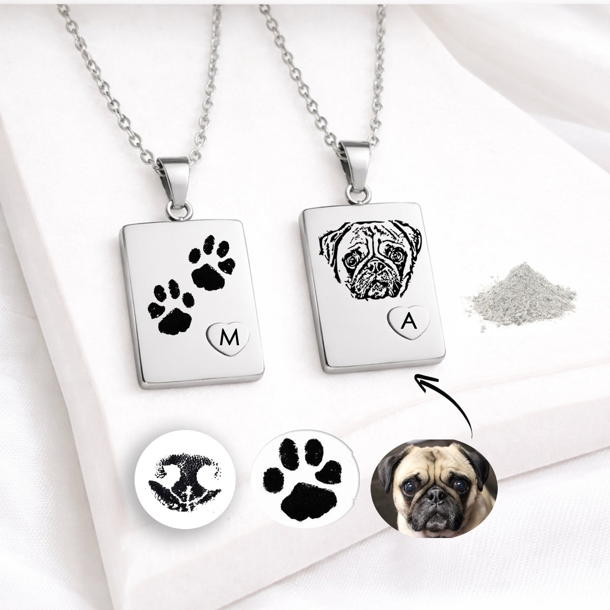 Two polished silver rectangular urn necklaces with matching chains on white fabric. One pendant is engraved with two black paw prints and a raised heart containing “M.” The other features an engraved dog portrait and a heart with “A.” Inset images show a nose print, paw print and pug photograph, with a small pile of ashes nearby.