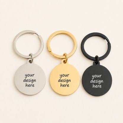Three round stainless steel keychains displayed in silver, gold, and black finishes on a warm ivory background. Each keychain has a smooth circular pendant engraved with “your design here” and is attached to a matching round key ring.