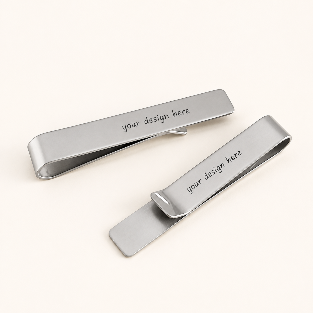 Two polished silver-tone rectangular tie clips displayed diagonally on a warm ivory background. Each stainless steel tie clip features rounded ends and the black personalized text “your design here” across the front.