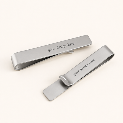 Two polished silver-tone rectangular tie clips displayed diagonally on a warm ivory background. Each stainless steel tie clip features rounded ends and the black personalized text “your design here” across the front.