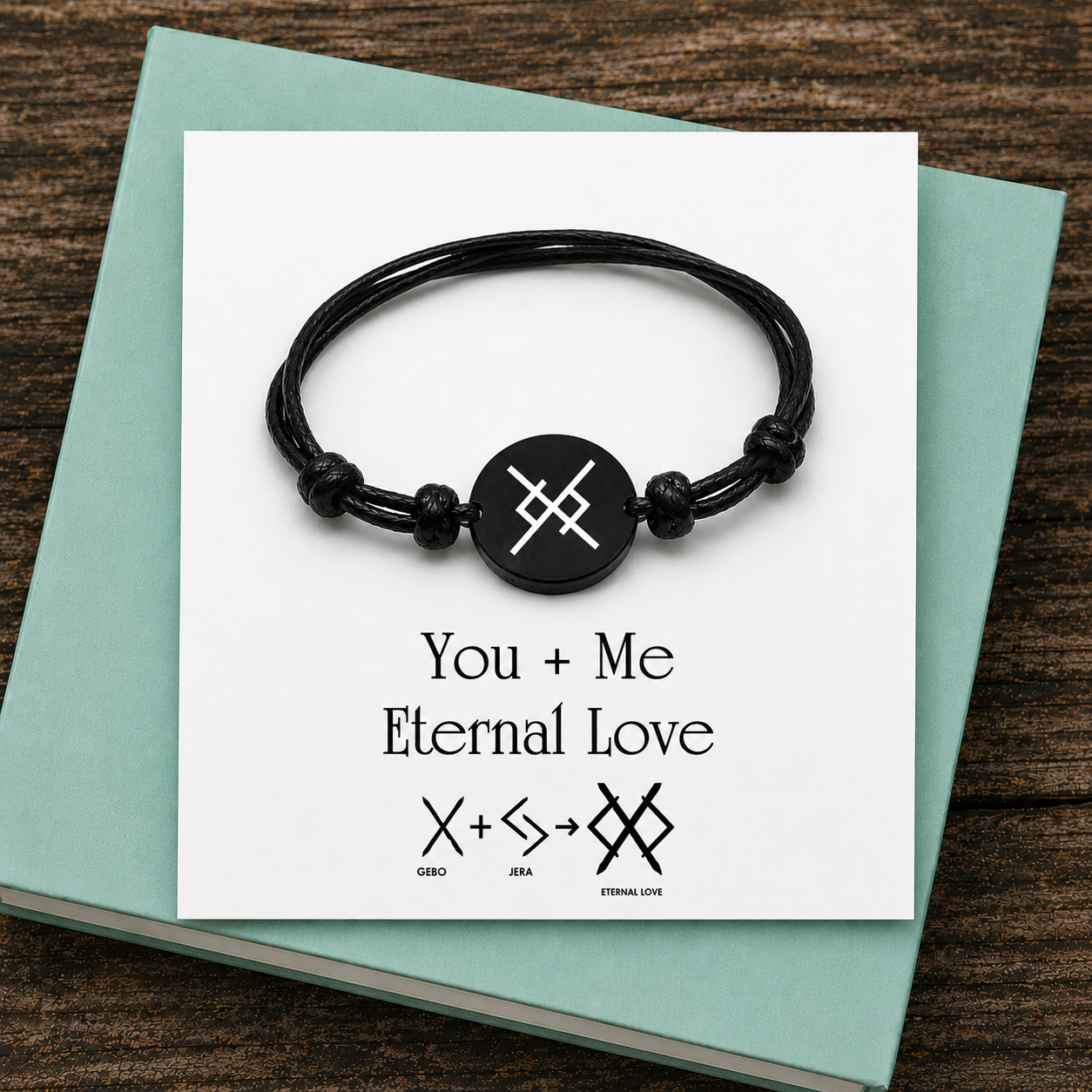 A black cord bracelet featuring a circular charm with a white runic symbol. The display card reads: You + Me Eternal Love, with the Gebo and Jera runes combined to form the Eternal Love symbol. The design is printed in black on white.